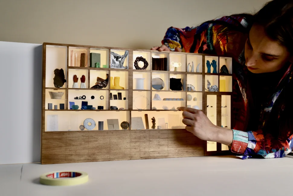 Modelmaker Maz stands to the right-hand side of a tiny 1:1 version of our Bloomsbury Materials Library cabinet. She is touching one of the materials on the shelves. On the shelves are miniature versions of our materials, including gloves, wellies, string, jars and material cubes.