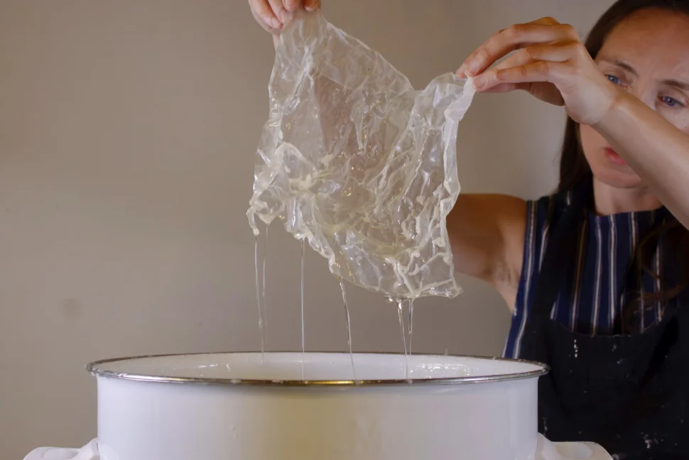 Yesenia stands to the right, holding a piece of white, slightly see-through paper above a large, white wax pot after dipping it in the liquid material. Yellowish wax is dripping from the crinkled material.
