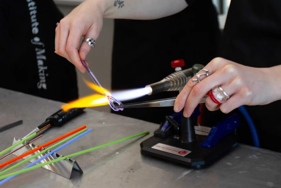 A close-up image of Roslyn lampnoodeling. She holds a piece of purple-tinged glass rod in a lamp's flame, using one hand to shape the piece with metal tweezers. To the left of the image are six pieces of colourful orange, green and blue glass.