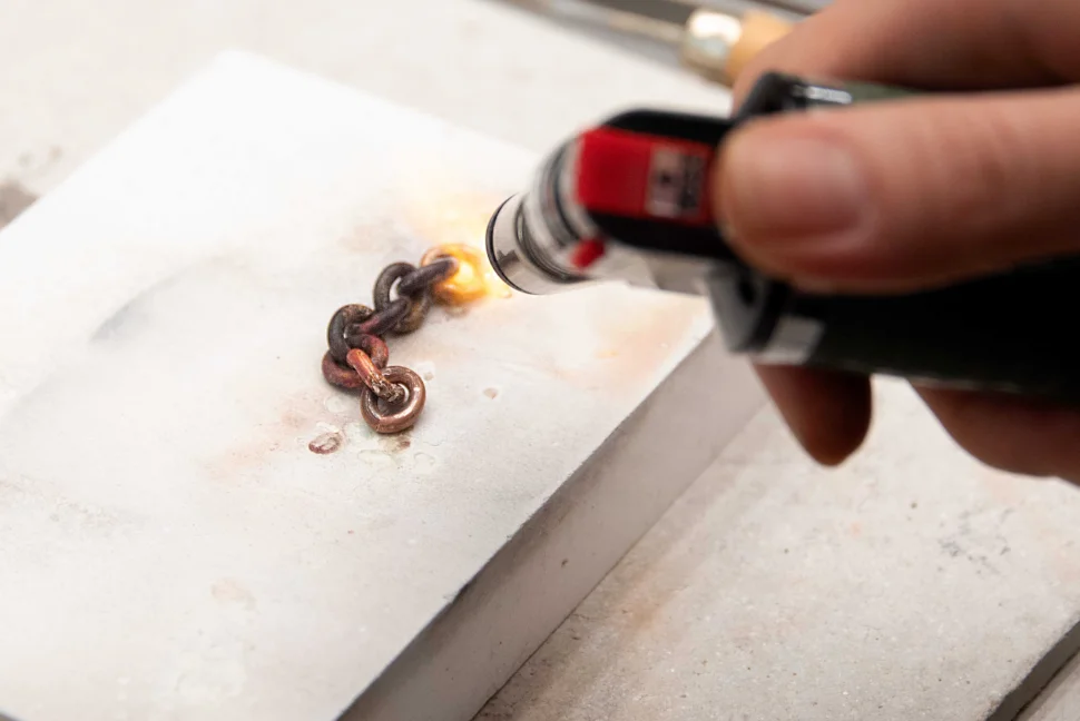 A close-up of someone using a torch to heat eight links of curb chain, on top of a grey slab. Someone is holding the torch to the right of the image, the flame hitting a piece of curb to the right of the link.