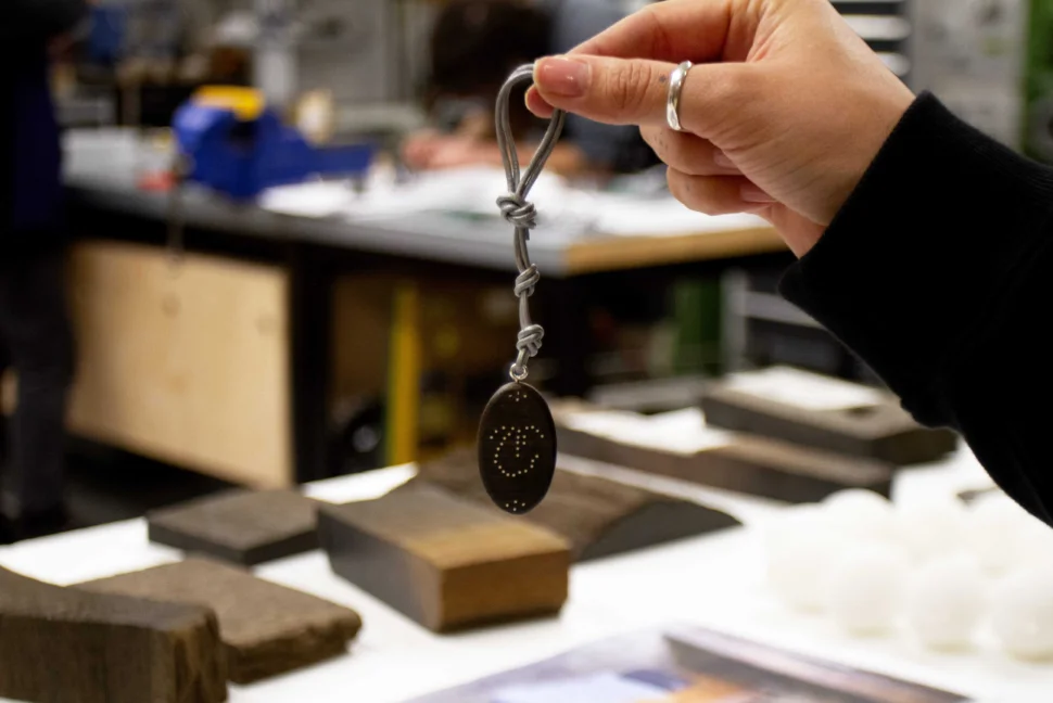 In the foreground, someone is holding a piece of bog oak that has been made into a pendant. The pendant is on a thick silver wire, and has an embellished design on the front in silver. The person holding the piece is wearing a silver ring and black long sleeve. In the background, you can see the Bloomsbury workshop, an array of bog oak sitting on top of the worktables.