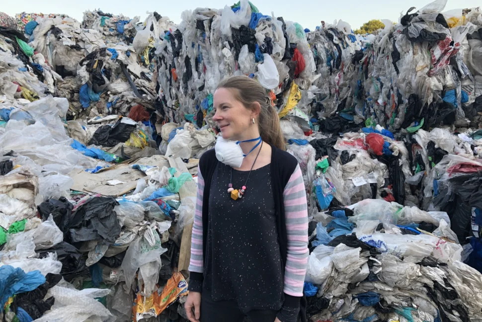 Sophie Thomas OBE stands at the forefront of a pile of plastic waste. She looks to her left side, a face mask hanging round her neck.