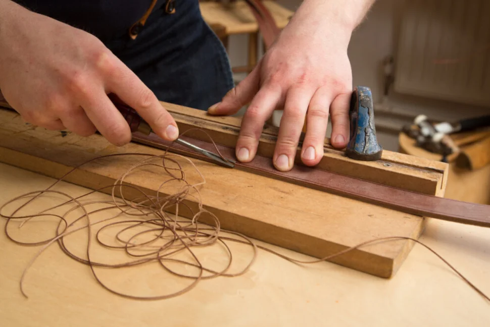 A person's hands are bevelling the edge of a piece of leather that is wedged against a block of wood. Strings of leather have been shaved off of the piece, and are lying to the side of the brown leather.
