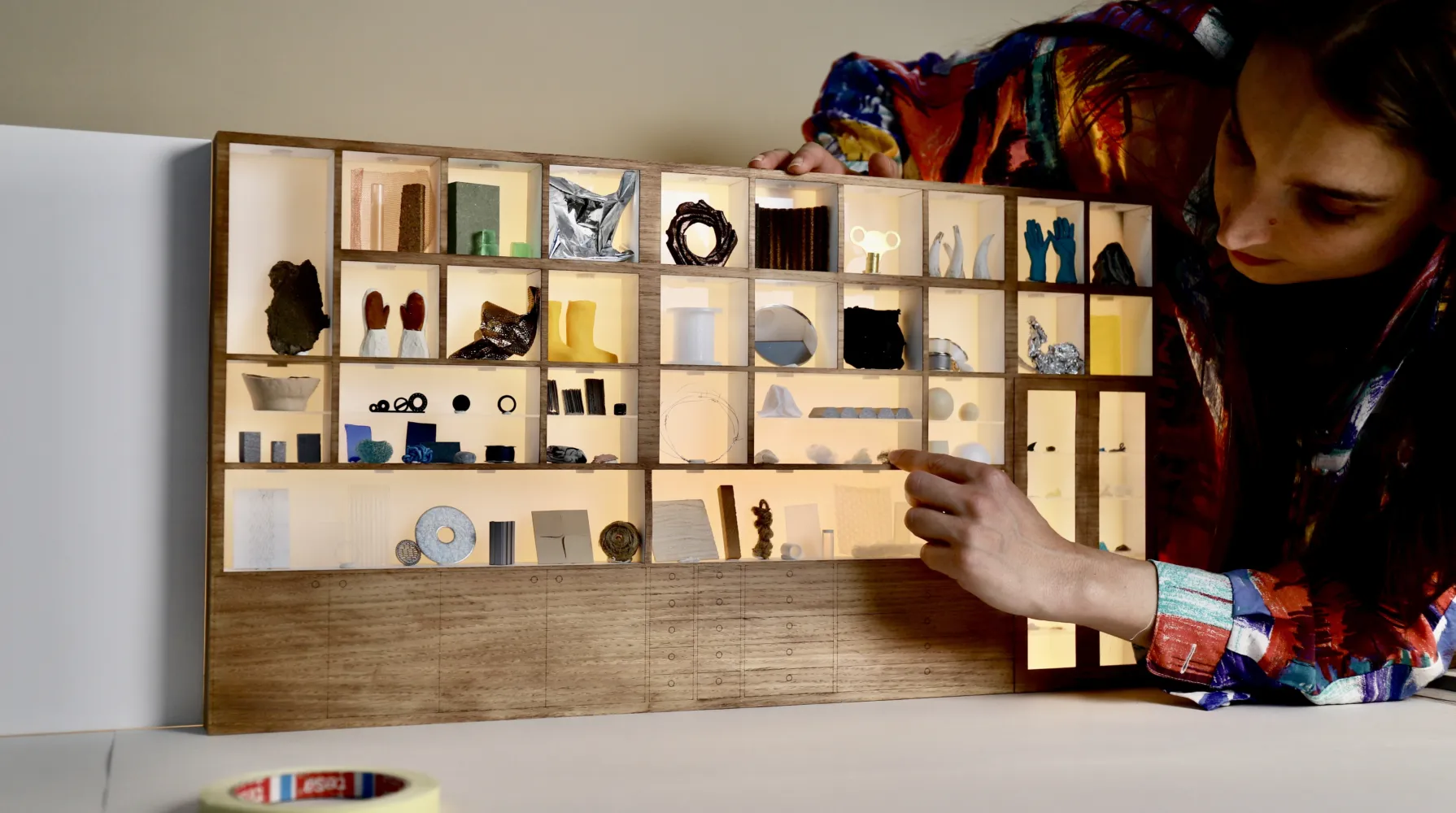 Modelmaker Maz stands to the right-hand side of a tiny 1:1 version of our Bloomsbury Materials Library cabinet. She is touching one of the materials on the shelves. On the shelves are miniature versions of our materials, including gloves, wellies, string, jars and material cubes.