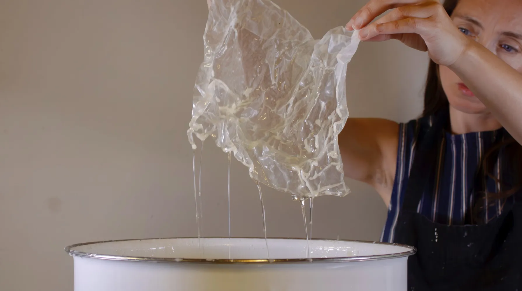 Yesenia stands to the right, holding a piece of white, slightly see-through paper above a large, white wax pot after dipping it in the liquid material. Yellowish wax is dripping from the crinkled material.