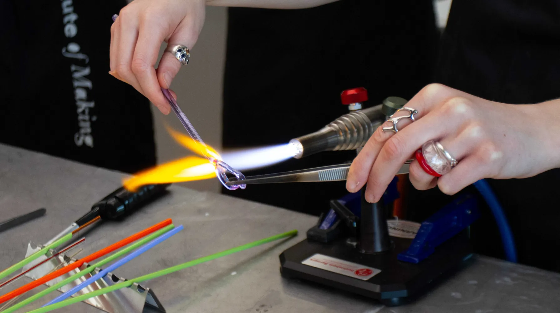 A close-up image of Roslyn lampnoodeling. She holds a piece of purple-tinged glass rod in a lamp's flame, using one hand to shape the piece with metal tweezers. To the left of the image are six pieces of colourful orange, green and blue glass.