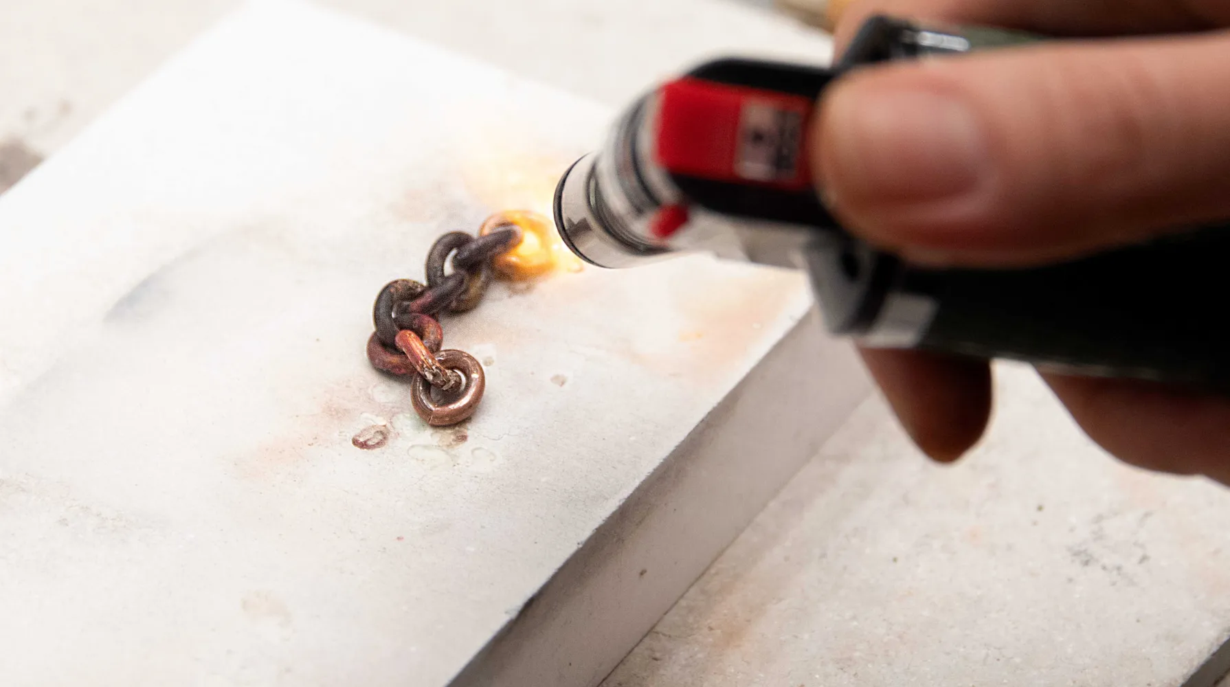 A close-up of someone using a torch to heat eight links of curb chain, on top of a grey slab. Someone is holding the torch to the right of the image, the flame hitting a piece of curb to the right of the link.