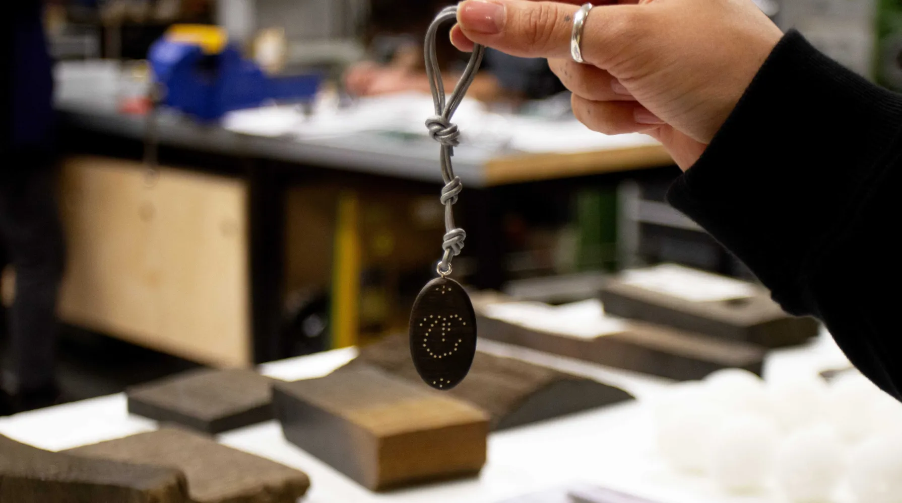 In the foreground, someone is holding a piece of bog oak that has been made into a pendant. The pendant is on a thick silver wire, and has an embellished design on the front in silver. The person holding the piece is wearing a silver ring and black long sleeve. In the background, you can see the Bloomsbury workshop, an array of bog oak sitting on top of the worktables.