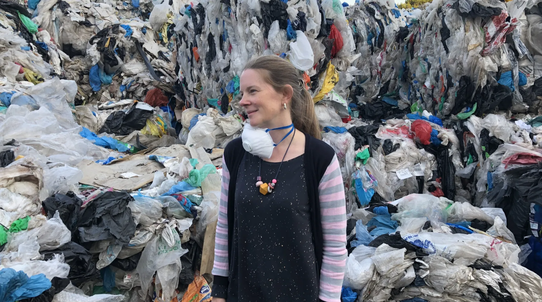 Sophie Thomas OBE stands at the forefront of a pile of plastic waste. She looks to her left side, a face mask hanging round her neck.