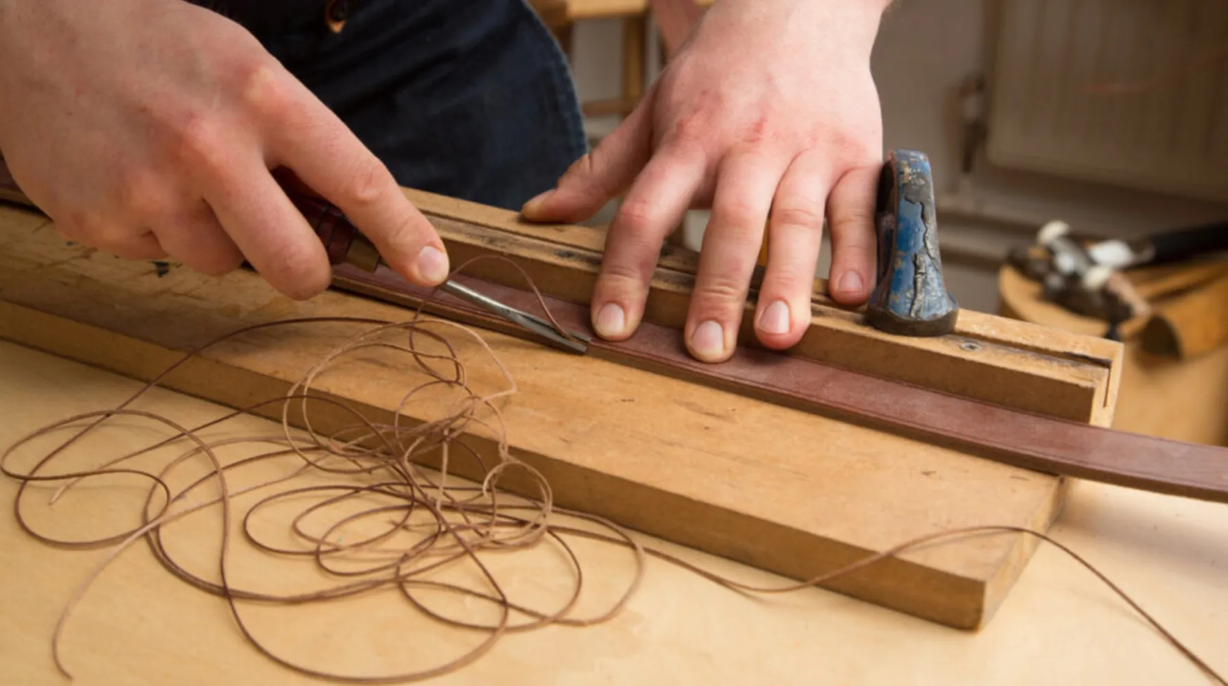 A person's hands are bevelling the edge of a piece of leather that is wedged against a block of wood. Strings of leather have been shaved off of the piece, and are lying to the side of the brown leather.