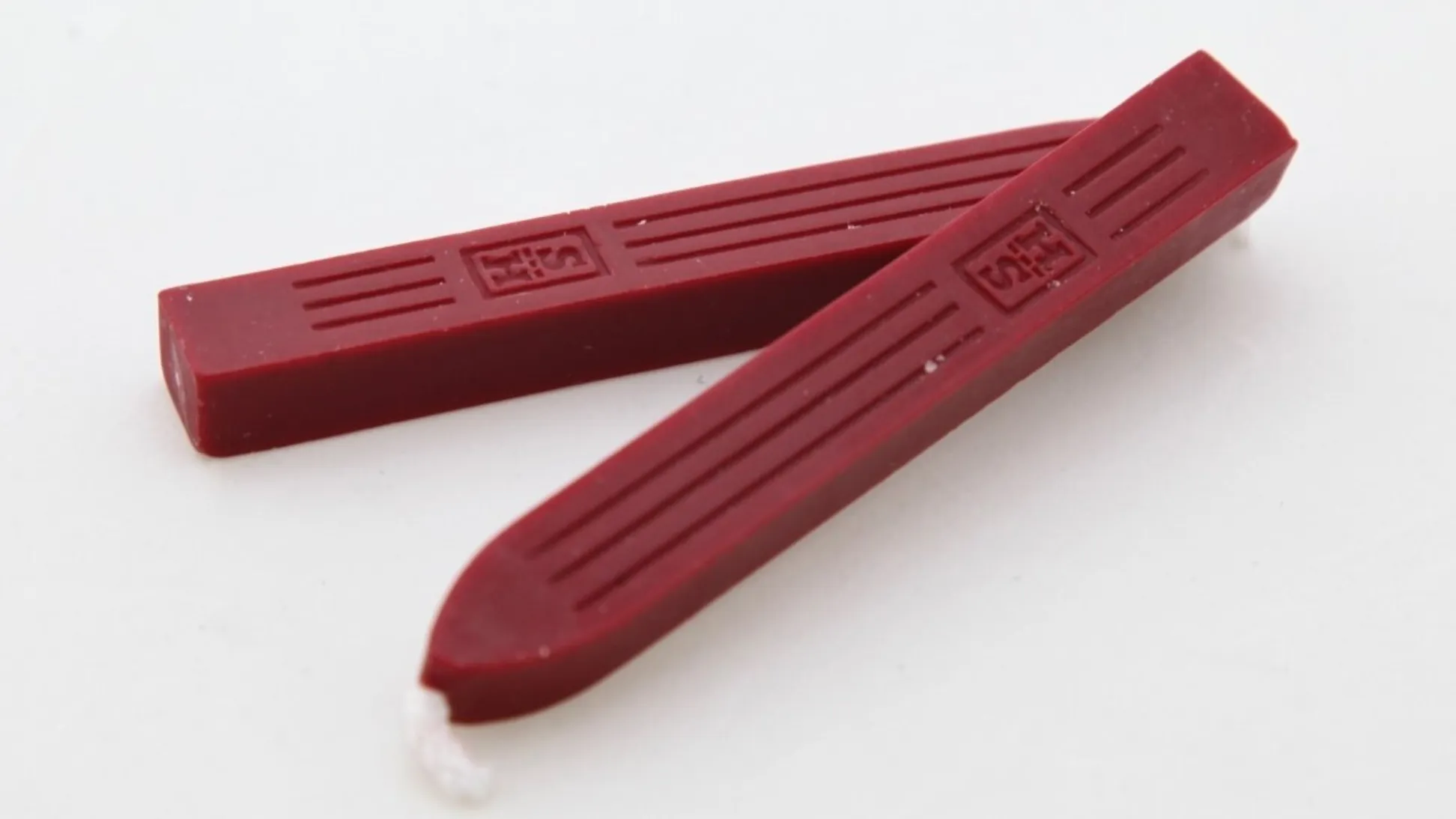 Two unused dark red coloured wax square edged candles laying on top of each other on a white surface